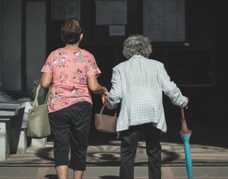 Deux femmes marchent côte à côte sur un trottoir, l'une aidant l'autre à porter ses sacs